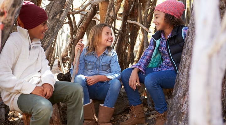 Three children sitting in a woodland den they’ve built.