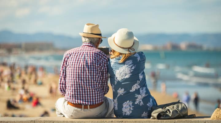 An older couple sitting on a wall at a beach.