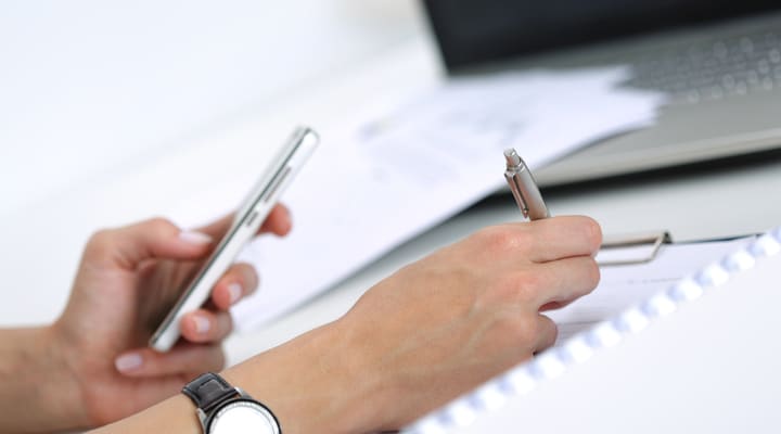 A person taking notes while looking at a phone screen.