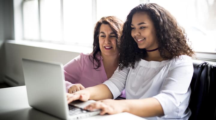 A happy mother and daughter sit at a desk working on a laptop together.