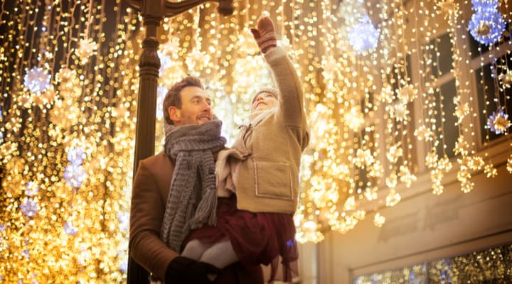 A father holds his young daughter in his arms as she reaches out towards hanging Christmas lights