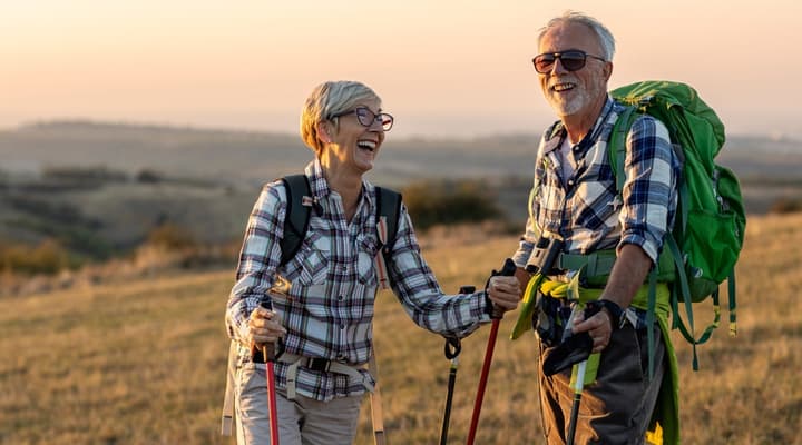 A smiling senior couple carry large backpacks across hilly terrain